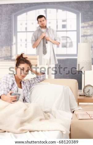 Young attractive woman drinking tea in bed in the morning, man dressing up in the background.