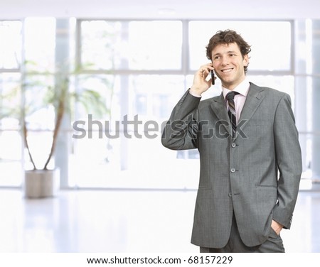 Young businessman standing in office lobby with hand in pocket, talking on mobile phone, smiling.?