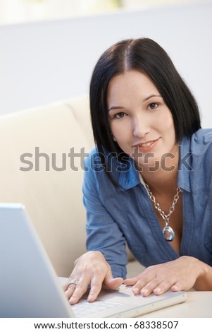 Portrait of young woman lying on couch with laptop computer, smiling at camera.