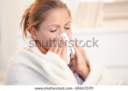 Young woman having flu, feeling bad, blowing her nose, wrapped up in blanket.?
