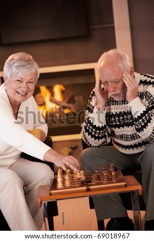 Couple playing chess in cosy living room in front of fireplace, elderly woman winning the game, senior man looking troubled.?