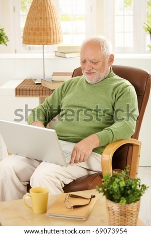 Smiling older man sitting in armchair using laptop computer at home.