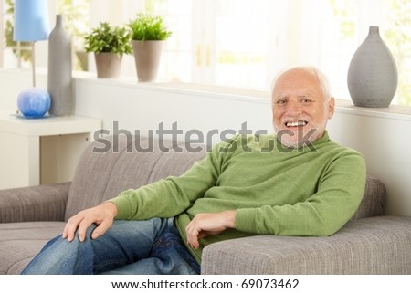 Portrait of happy senior man sitting on living room sofa, laughing, looking at camera.