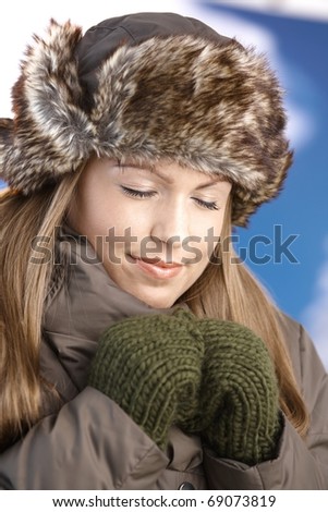Young attractive female dressed up warm in coat, fur-hat and gloves, enjoying winter sun, smiling, eyes closed.