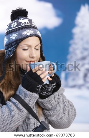 Pretty young girl dressed up warm for skiing wearing cap and gloves drinking hot tea eyes closed front of winter landscape .