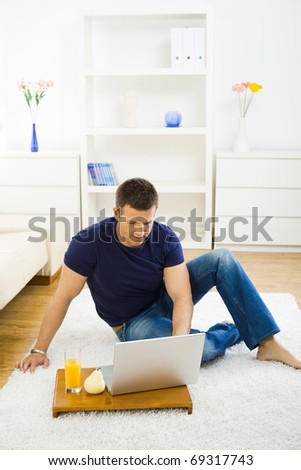 Casual young man working at home on his laptop computer, sitting on floor looking at screen.?