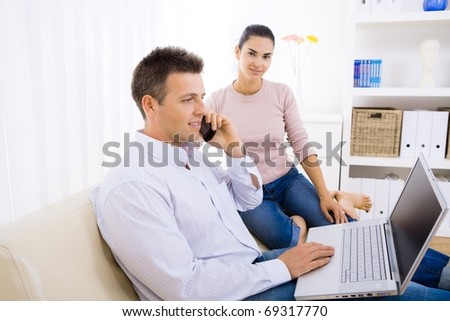 Young couple using laptop computer at home, sitting on couch. Man talkin on mobile phone. Selective focus on man.?