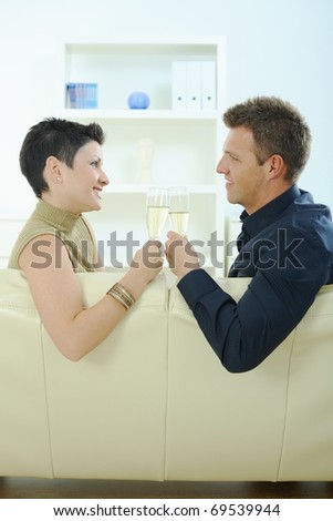 Love couple clinking champagne glasses at home on sofa. Smiling and looking at each other.