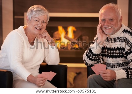 Portrait of happy senior couple playing cards at home in front of cosy fireplace, smiling at camera.?