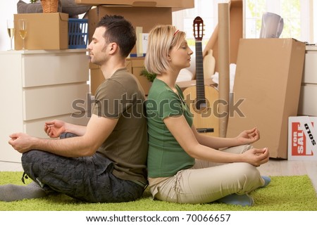 Young couple sitting in yoga meditation posture on carpet in new house, surrounded with boxes.?