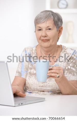 Senior woman sitting at desk using laptop computer, drinking tea.?