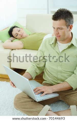 Man sitting on floor at home browsing internet on laptop computer, woman sleeping on sofa.?