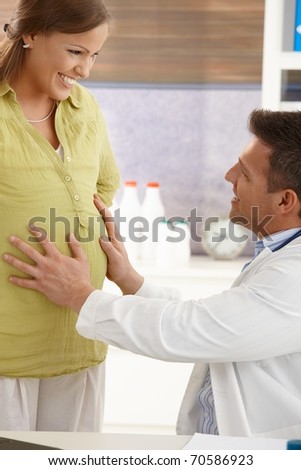 Smiling doctor touching belly of happy pregnant woman at examination in consulting room.?