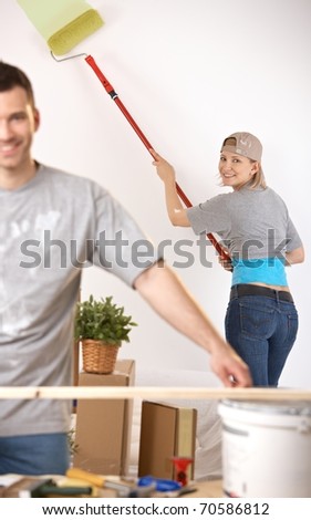 Smiling young woman in focus painting wall with paint roller, man working at table.?