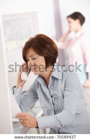 Mature office worker woman using landline phone, smiling, coworker using mobile phone in background.?