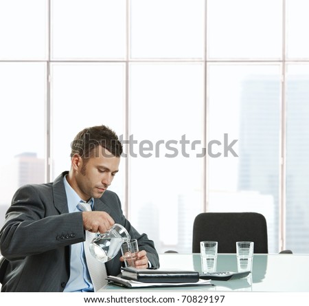 Thirsty businessman sitting at meeting table pouring water from jug into glass to drink in meeting break.