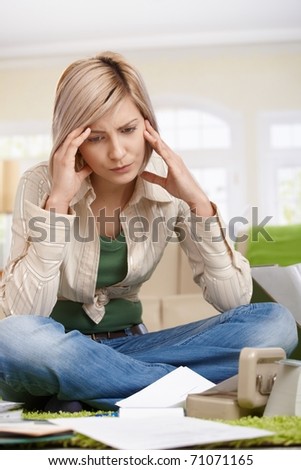 Troubled woman holding head sitting on living room floor looking at documents.?