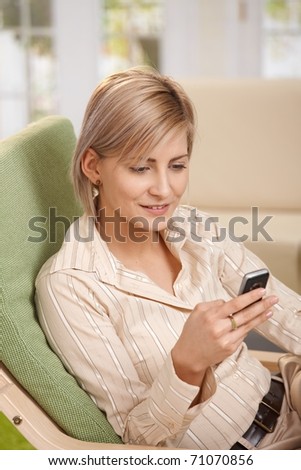 Smiling blonde woman sitting in armchair at home, using mobile phone, having coffee mug on table.?