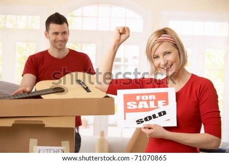 Smiling woman holding sold sign raising fist, happy man looking at her in background, unpacking boxes.?