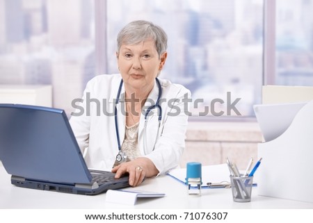 Senior female doctor, working at desk, using laptop computer. Looking at camera.?