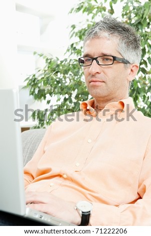Mid-adult man wearing jeans and orange shirt sitting on couch, working with laptop computer. ?
