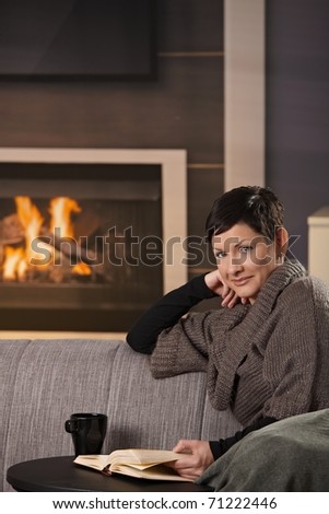 Woman sitting on sofa at home on a cold winter day, reading book, looking up at camera.?