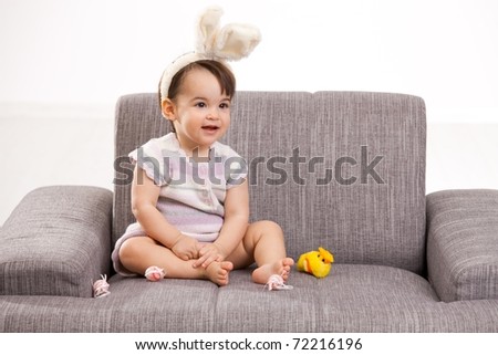 Baby girl in easter bunny costume, sitting on grey couch playing with toy chicken and easter eggs, laughing. Isolated on white background.?