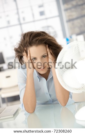 Young attractive woman sitting in office front of fan, feeling hot, cooling herself.?