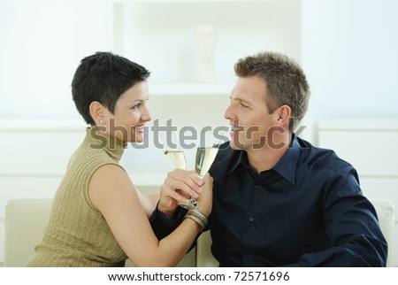 Love couple clinking champagne glasses at home on sofa. Smiling and looking at each other.?