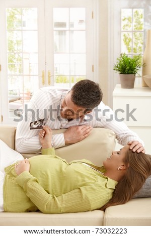 Expecting woman lying on sofa, man stroking her hair, looking at ultrasound baby photo together.?