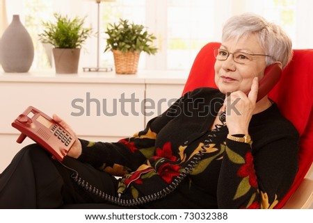 Pensioner woman using landline phone sitting in armchair in living room.?