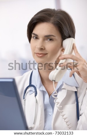 Mid-adult female doctor sitting behind computer and calling, looking at camera, smiling.?