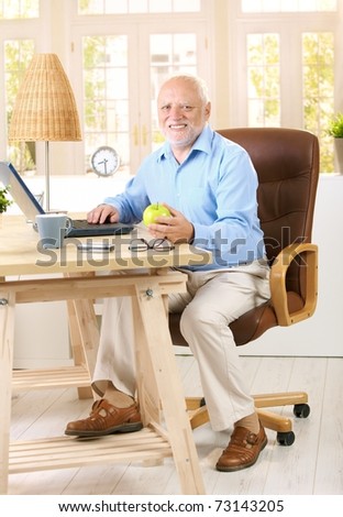 Older man working in his study at home, using computer, holding apple, looking at camera, smiling.?