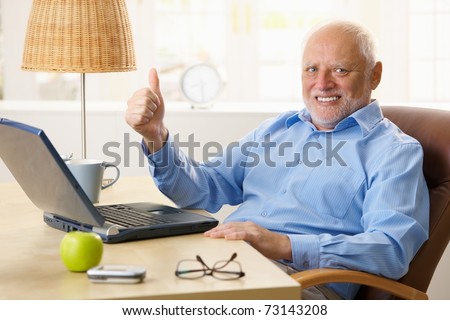 Happy senior man giving thumb up, sitting at desk using laptop computer at home.?