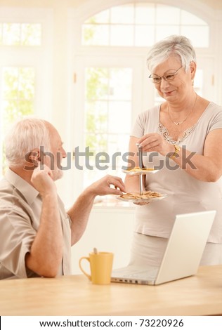 Smiling wife serving cookies in kitchen to husband sitting with laptop computer and coffee.?