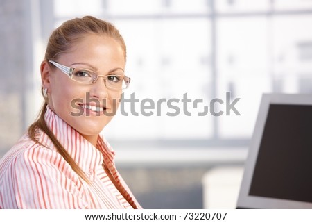 Portrait of young woman sitting at desk, working with computer, smiling.?