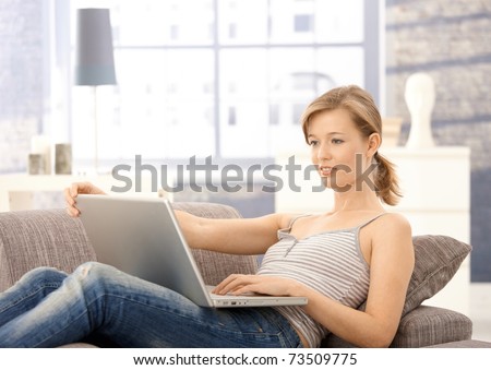 Young woman lying on sofa at home, browsing internet on laptop. Looking at screen, smiling.?