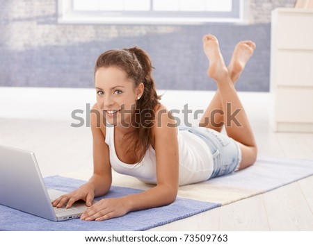 Attractive young female using laptop, downloading e-mails, laying on floor at home, smiling.