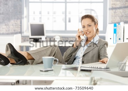Young attractive businesswoman sitting at desk, legs on top of desk, talking on phone, smiling.