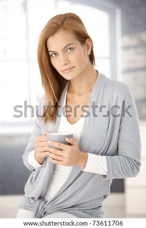 Portrait of beautiful young woman standing, having coffee in bright room.?