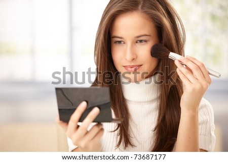Attractive woman applying makeup with brush, checking in pocket mirror, smiling.?