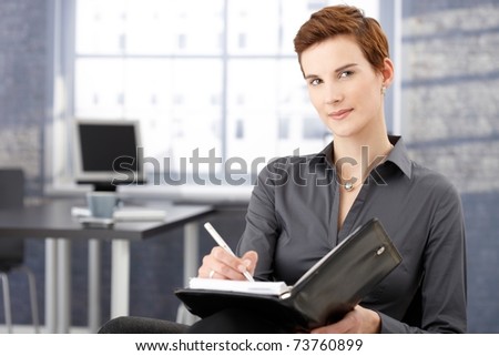 Smiling businesswoman writing notes in office, looking at camera, smiling.?