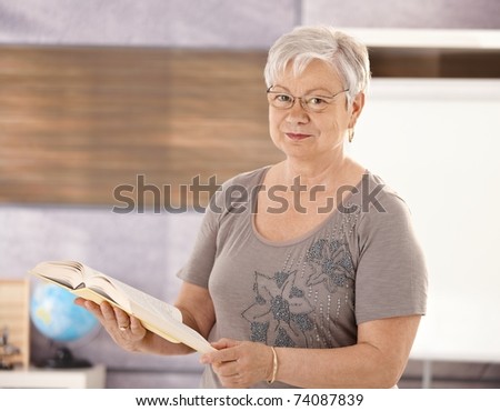 Portrait of senior teacher standing in classroom, holding book, looking at camera.?