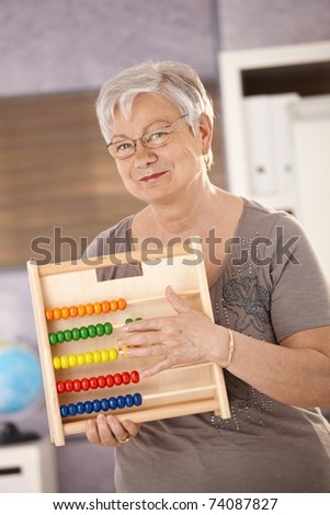 Senior teacher standing in classroom, holding abacus, smiling.?