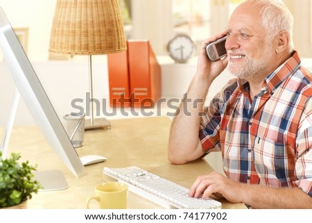 Senior man talking on cellphone, using computer, smiling, looking at screen at home.?