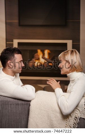Young romantic couple sitting on couch in front of fireplace at home, looking at each other, talking.?