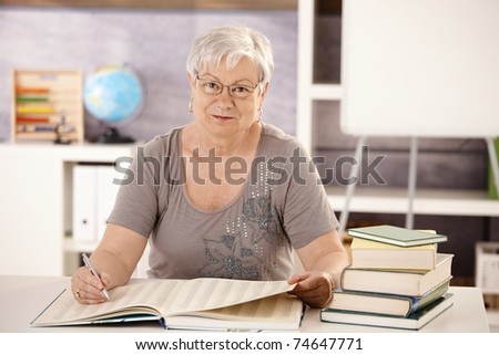 Portrait of senior teacher sitting at desk in classroom, searching in a book, looking at camera.?