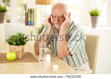 Senior man sitting at table, having bad headache, grimacing, taking medicine.?