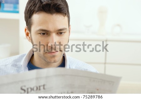 Handsome young man sitting on couch at home reading sport news in newspaper.?