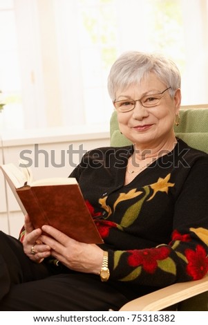 Portrait of elderly woman sitting in armchair with book in hand, smiling at camera.?